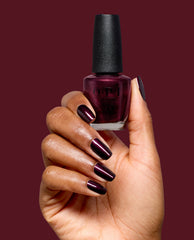 Hand holds OPI Every Month is Oktoberfest nail lacquer, showing deep burgundy shimmer polish on manicured nails against a wine-red background, ideal for fall nail colors.