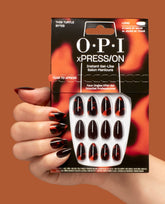 A hand wearing OPI xPRESS/ON This Turtle Bytes press-on nails holds the pack, showing long gel-like tortoiseshell black and burnt orange fall nail colors on a warm brown background.