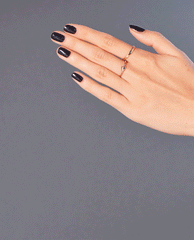Hand with short, glossy nails painted in Rub-a-Pub-Pub, a deep charcoal OPI nail polish nail lacquer, posed against a soft gray background with delicate gold rings to showcase the rich, opaque color and smooth salon-quality finish.