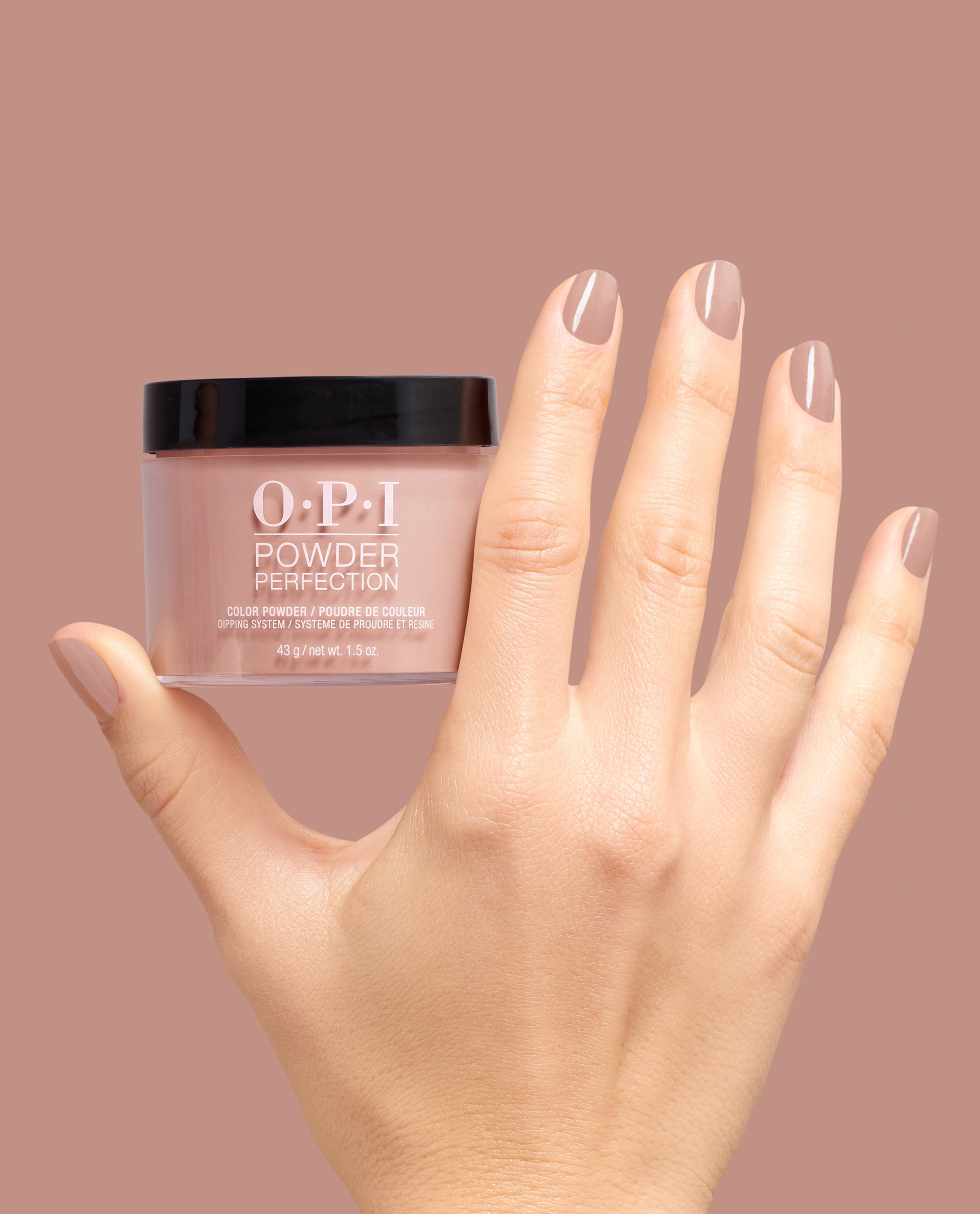 Hand holding OPI Powder Perfection France-y Seeing You Here dipping powder, with glossy taupe-mauve fall nail colors shown on manicured nails against a matching background.