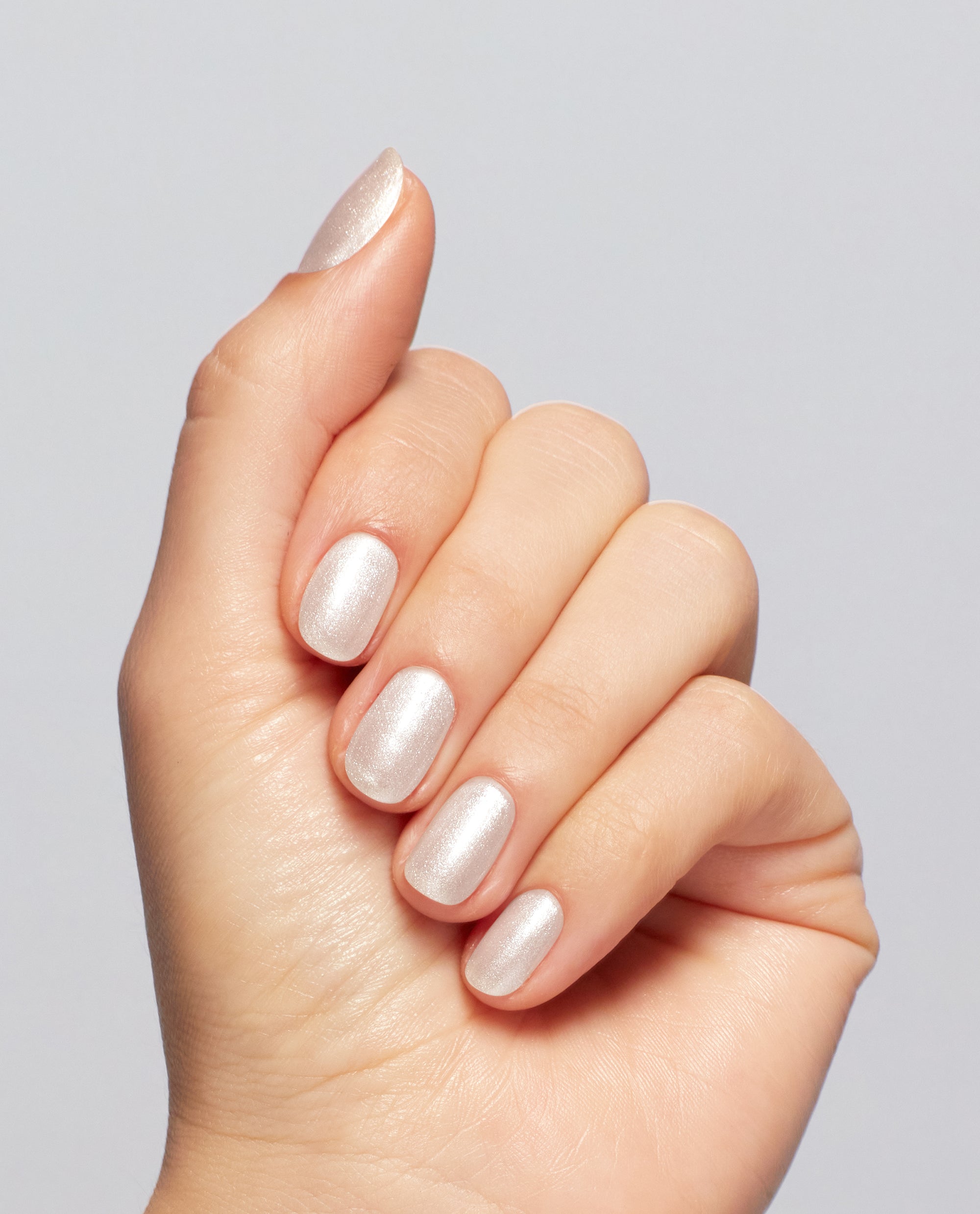 Hand showing short oval nails painted with Gemini and I nail lacquer, a shimmery pearlescent white OPI nail polish, against a soft light gray background, highlighting the smooth, glossy manicure finish.