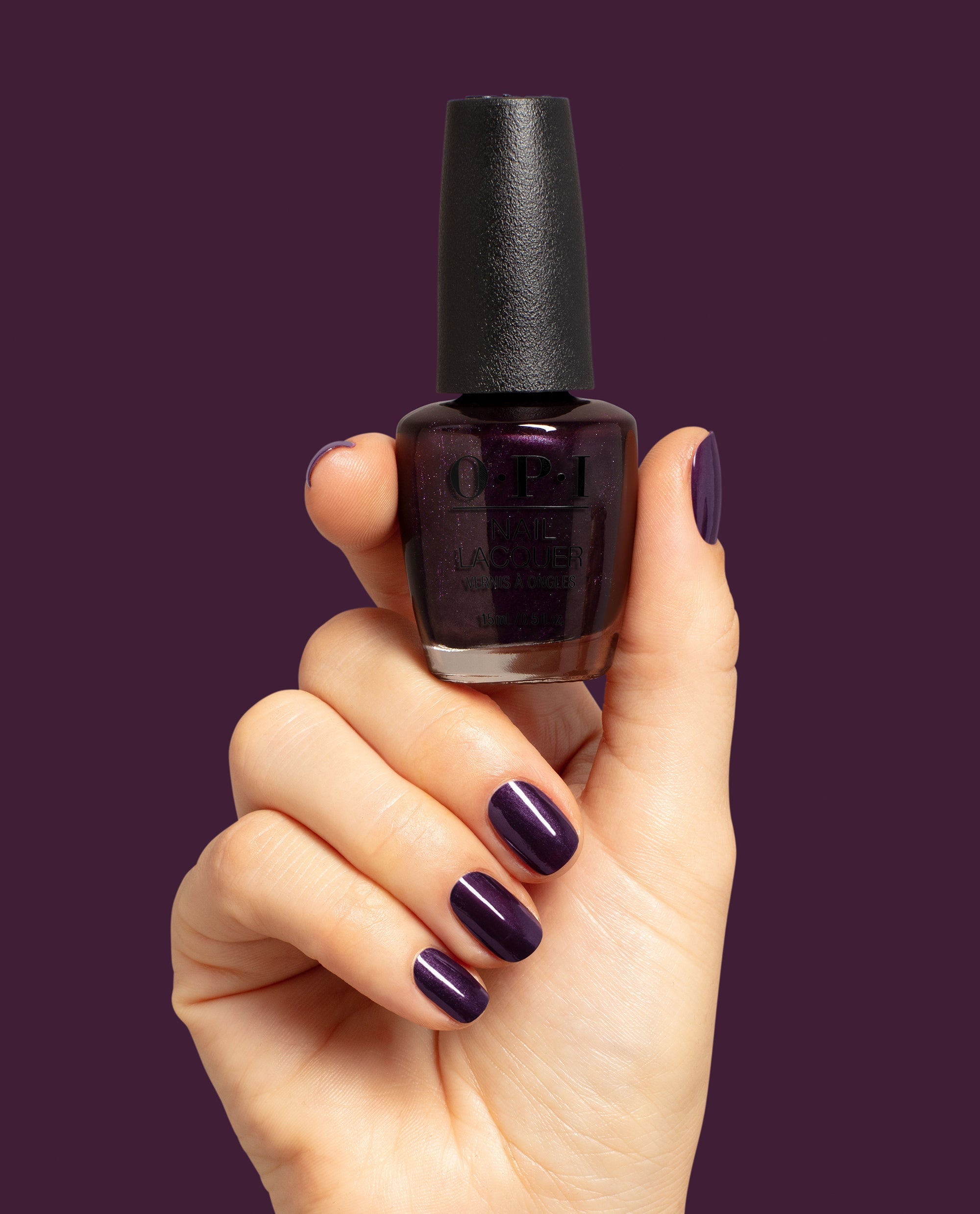 Hand with glossy deep purple nails holds OPI Lincoln Park at Midnight Nail Lacquer bottle against a plum background, showcasing a dark shimmer shade for fall nail colors.