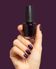 Hand with glossy deep purple nails holds OPI Lincoln Park at Midnight Nail Lacquer bottle against a plum background, showcasing a dark shimmer shade for fall nail colors.