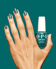 Manicured hand wearing shimmery deep teal green nail polish holds an OPI GelColor Yuletide Sweetings! gel nail polish bottle against a matching teal background, showcasing glossy sparkle; green nail polish pick for spring nail colors from OPI.